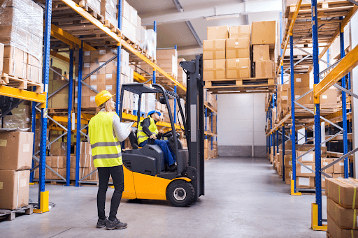 A man on a forklift being guided to add boxes on the top shelf